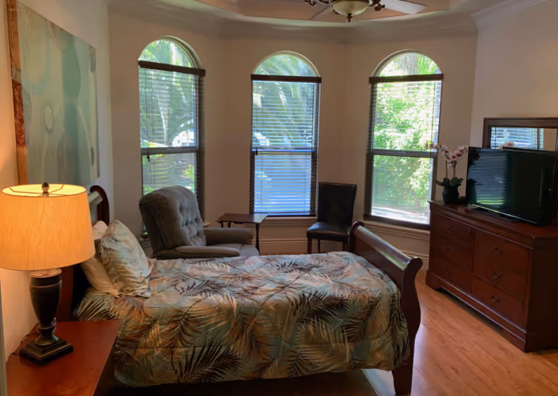 A cozy bedroom with a single bed covered in a leafy patterned bedspread. There is a wooden nightstand with a lamp on the left side of the bed. Next to the bed is a comfortable armchair and a small table. Three tall windows with blinds allow natural light to fill the room. On the right side, there is a wooden dresser with a flat-screen TV and a small potted orchid on top. The room has wooden flooring and light-colored walls.