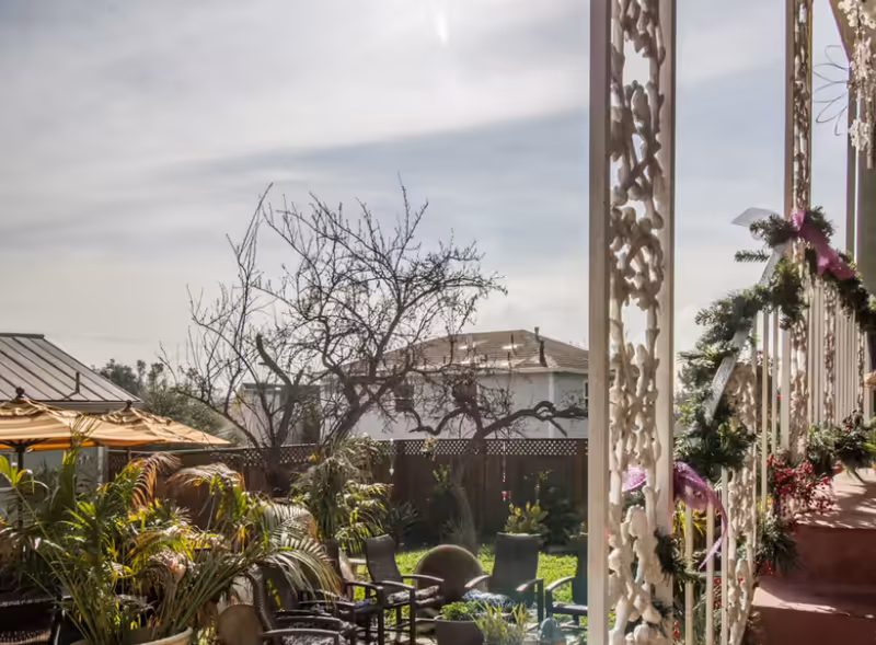 Outdoor patio area with several chairs and tables surrounded by various plants and greenery. A wooden fence and leafless tree are visible in the background under a partly cloudy sky. The right side shows a porch with decorative white railings adorned with garlands and ribbons.