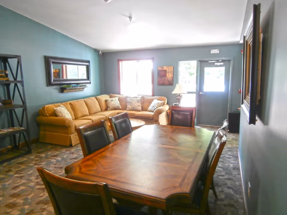 A cozy living room area with a large beige sectional sofa adorned with patterned throw pillows, a wooden dining table with six chairs in the foreground, a tall black bookshelf on the left wall, a large rectangular mirror above the sofa, and a door and window letting in natural light at the back.