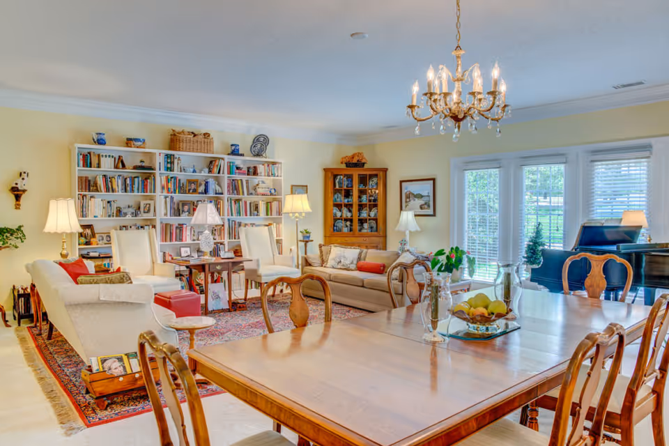 A bright and spacious living and dining area with a wooden dining table and chairs in the foreground. The living area features comfortable seating including sofas and armchairs, a large bookshelf filled with books and decorative items, a wooden cabinet, and several lamps providing warm lighting. Large windows and glass doors allow natural light to fill the room, and a chandelier hangs above the dining table. A grand piano is visible near the windows.