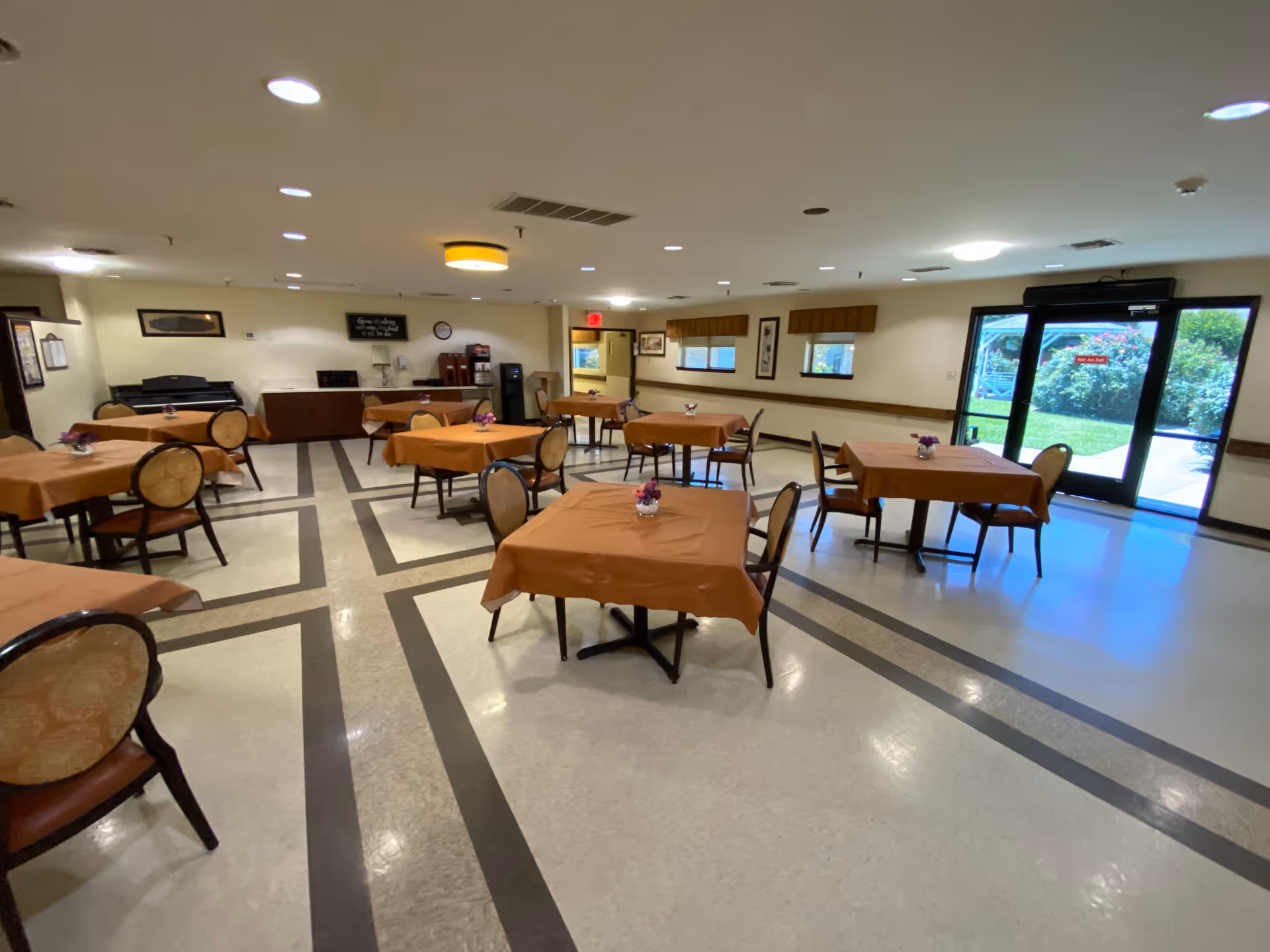 Spacious communal dining room with several tables covered in orange tablecloths, chairs, and glass doors opening to an outdoor area.
