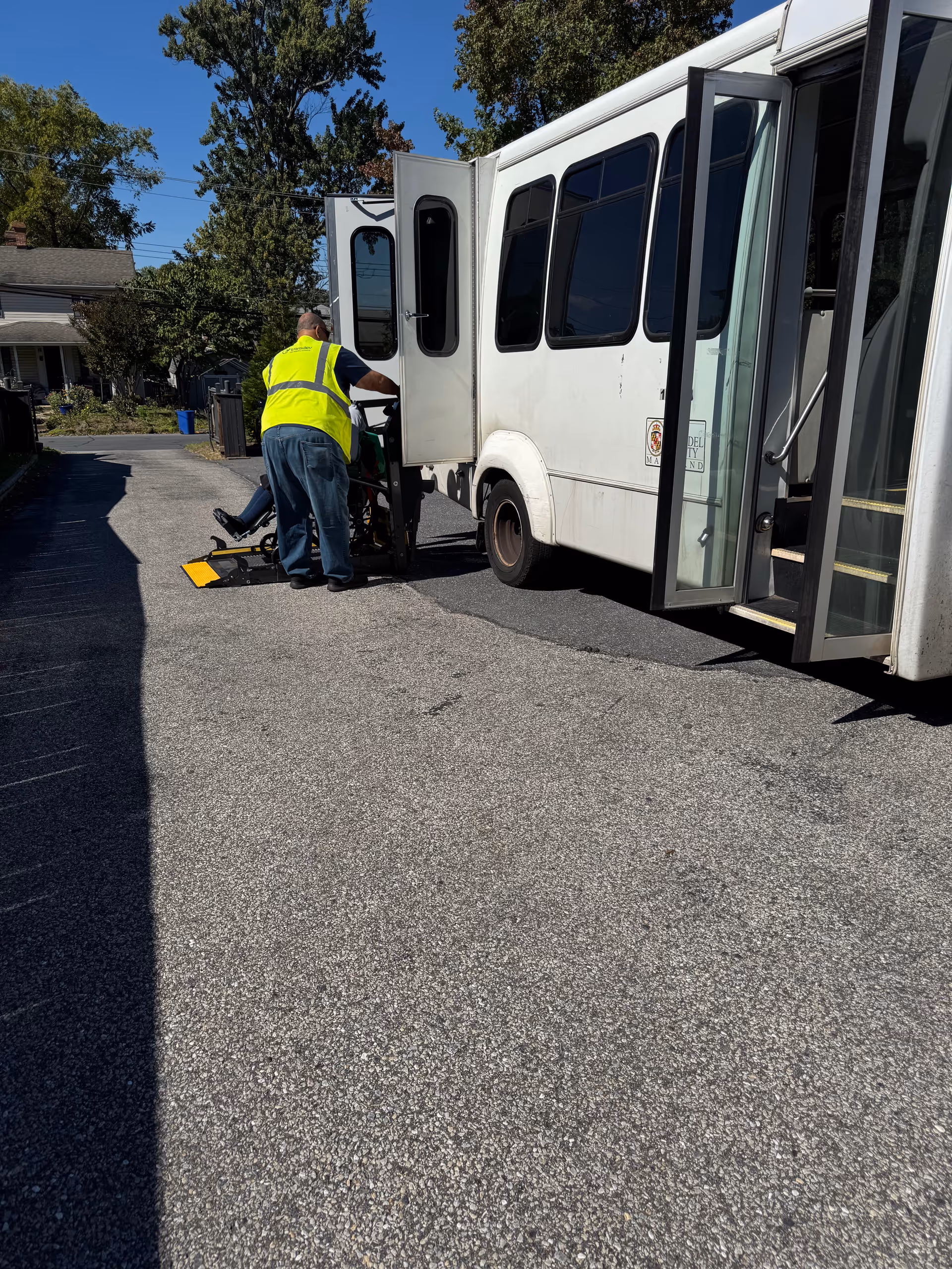 A worker in a yellow safety vest helps a person in a wheelchair onto a lift at the open door of a white shuttle bus parked on a residential street.