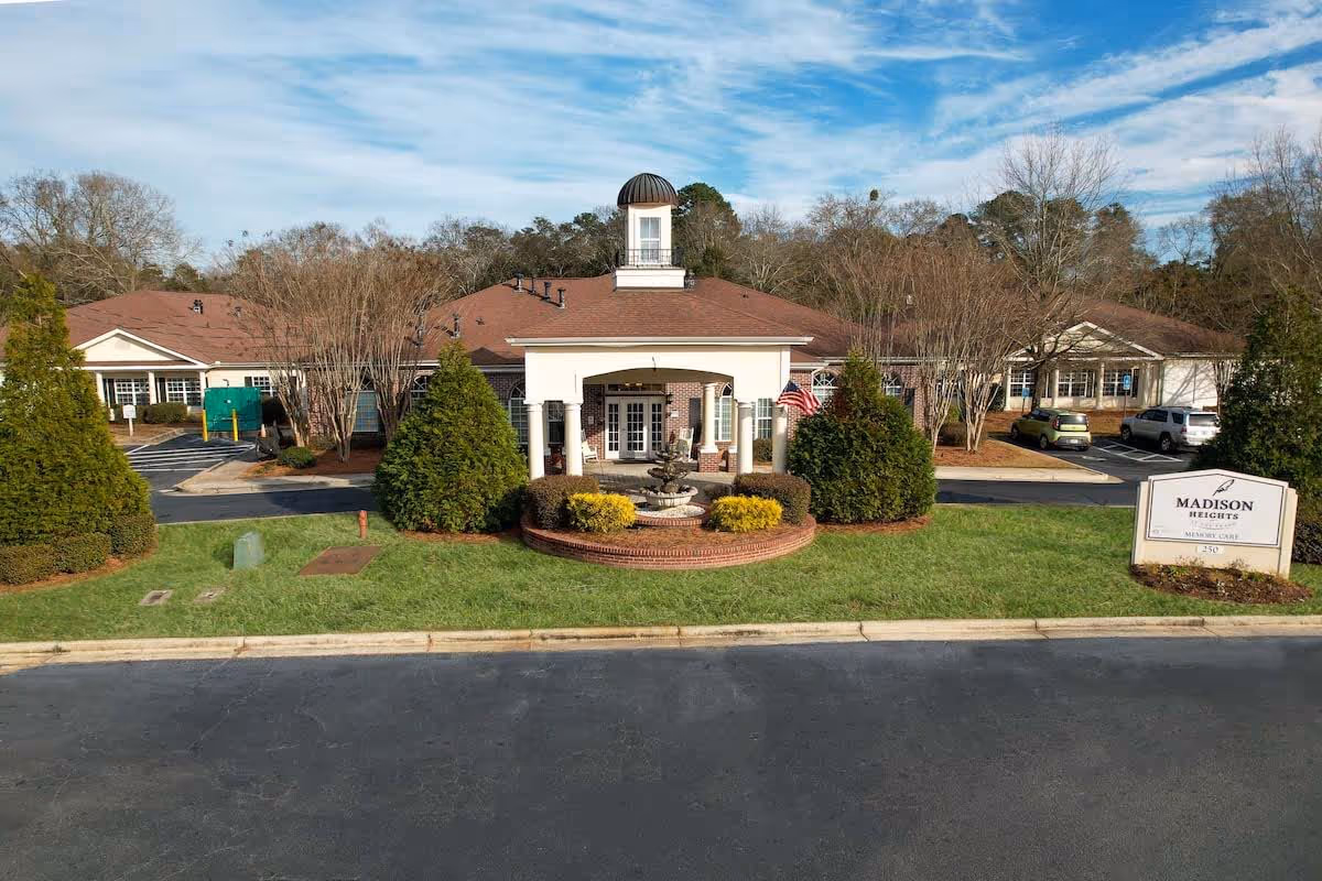 Front exterior of a single-story senior living building with a cupola, central entrance, fountain, and landscaped lawn.