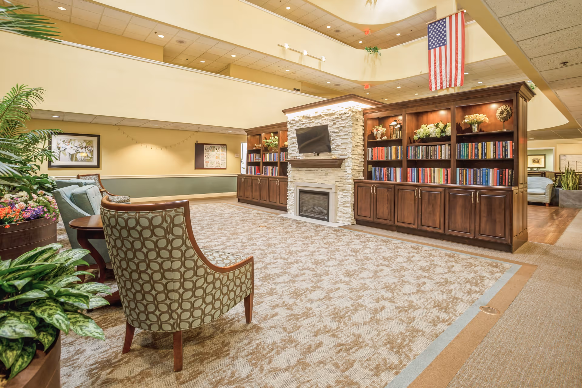 A spacious and well-lit common area with patterned armchairs and a side table in the foreground. The room features a large built-in wooden bookshelf filled with books and decorative flowers, centered around a stone fireplace with a mounted flat-screen TV above it. An American flag hangs from the ceiling, and there are plants and framed artwork on the walls. The carpet has a beige and light brown pattern, and the ceiling has recessed lighting.