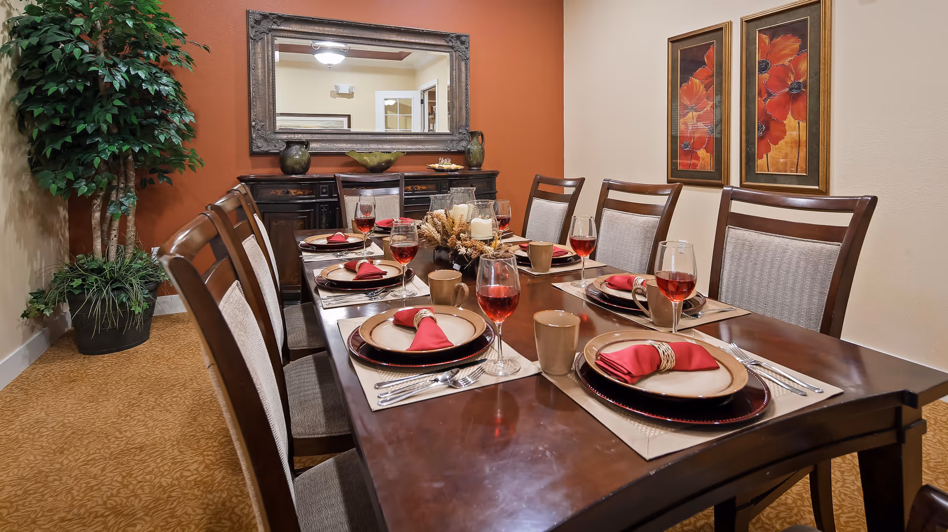 A formal dining room with a dark wooden table set for six people. Each place setting includes a plate with a red napkin, a wine glass filled with red liquid, and a beige mug. The room features a large mirror on a burnt orange accent wall, two framed floral paintings, a sideboard with decorative items, and a large potted plant in the corner.