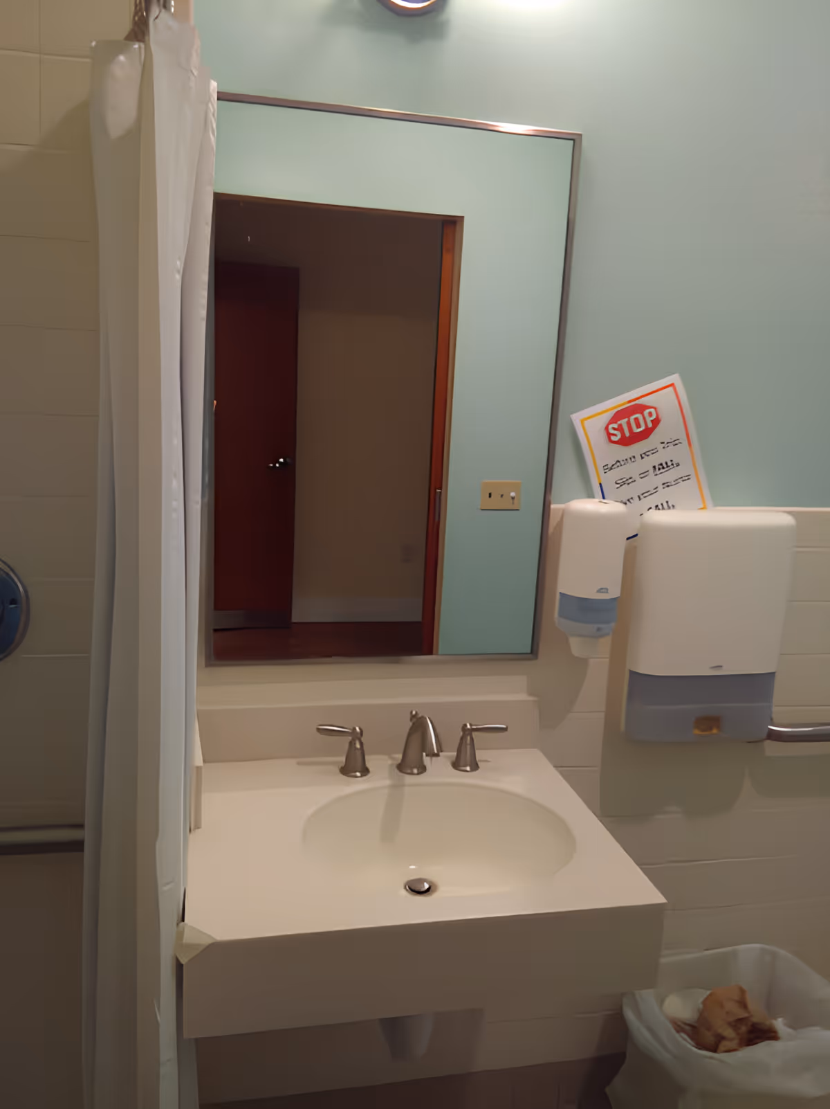 A bathroom sink with a silver faucet and two handles, a large rectangular mirror above it, a soap dispenser and paper towel dispenser mounted on the wall to the right, a shower curtain partially visible on the left, and a trash bin below the dispensers containing some used paper towels. A STOP sign notice is partially visible behind the dispensers.
