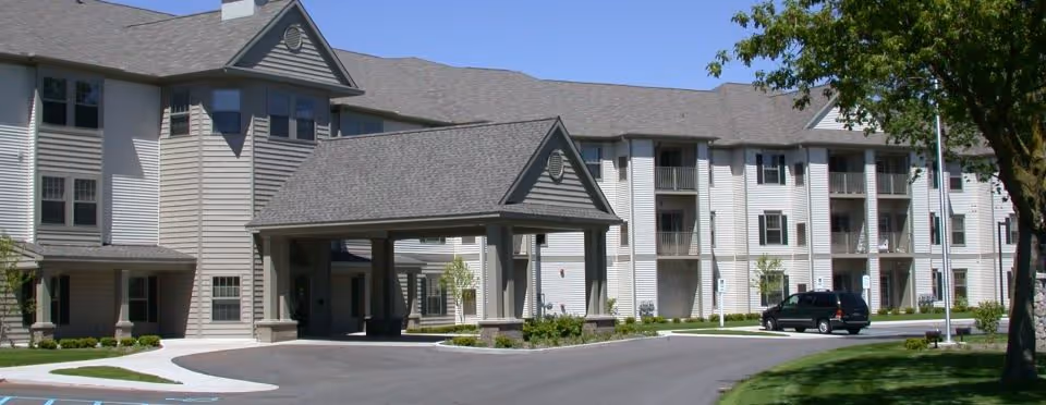 Exterior front view of the Village at Park Terrace building showing a covered porte-cochère, three-story siding with balconies, and a driveway with a parked vehicle.
