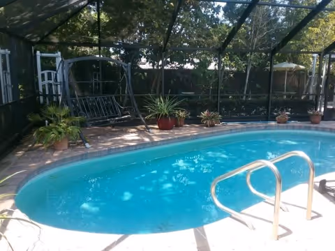 Outdoor swimming pool enclosed by a screened structure with several potted plants around the pool deck and a metal pool ladder in the foreground.