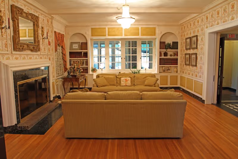 A cozy living room with two beige sofas facing a fireplace with a large mirror above it. The room has wooden floors, floral wallpaper, built-in bookshelves with books and decorative items, and a window with multiple panes letting in natural light. A chandelier hangs from the ceiling.
