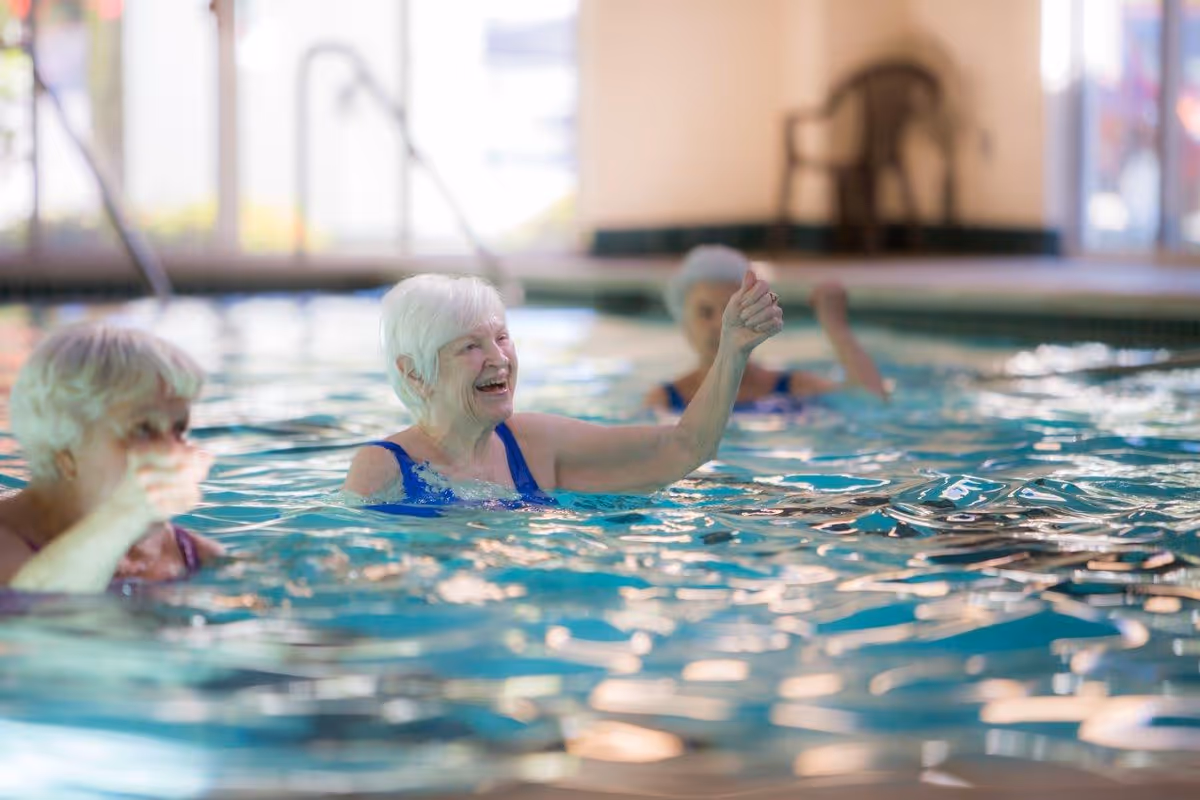 Three older adults participating in a water exercise class in an indoor pool, one smiling and raising her arm.