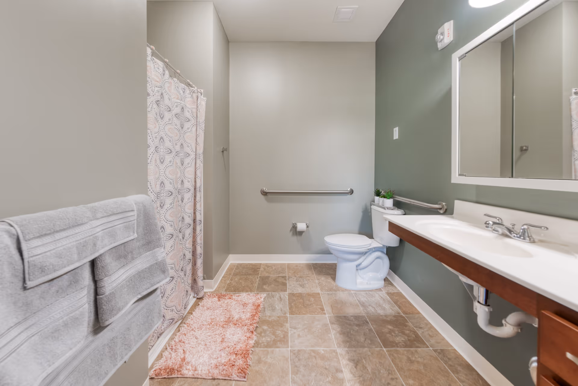 A clean and spacious bathroom with beige tiled floor, a white toilet with a grab bar beside it, a white sink with a wooden cabinet underneath, a large mirror above the sink, a shower with a patterned curtain, and two gray towels hanging on a towel rack. There is also a pink bath mat on the floor and a small plant on the toilet tank.