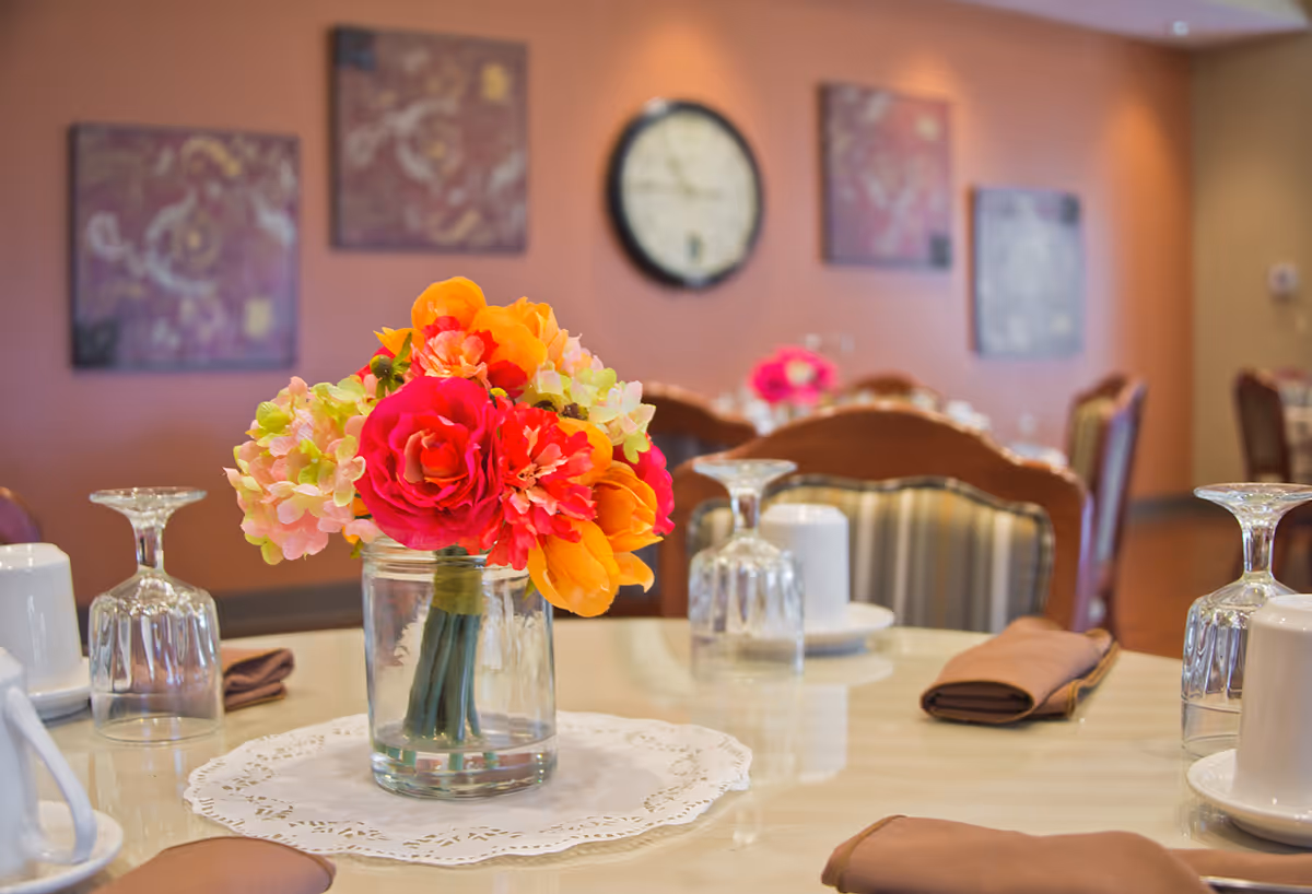 A dining table set with upside-down wine glasses, white cups on saucers, and brown folded napkins. A glass jar with a colorful bouquet of artificial flowers in shades of pink, orange, and green is placed in the center on a white doily. In the background, there are more tables and chairs, a wall clock, and decorative wall art on a peach-colored wall.
