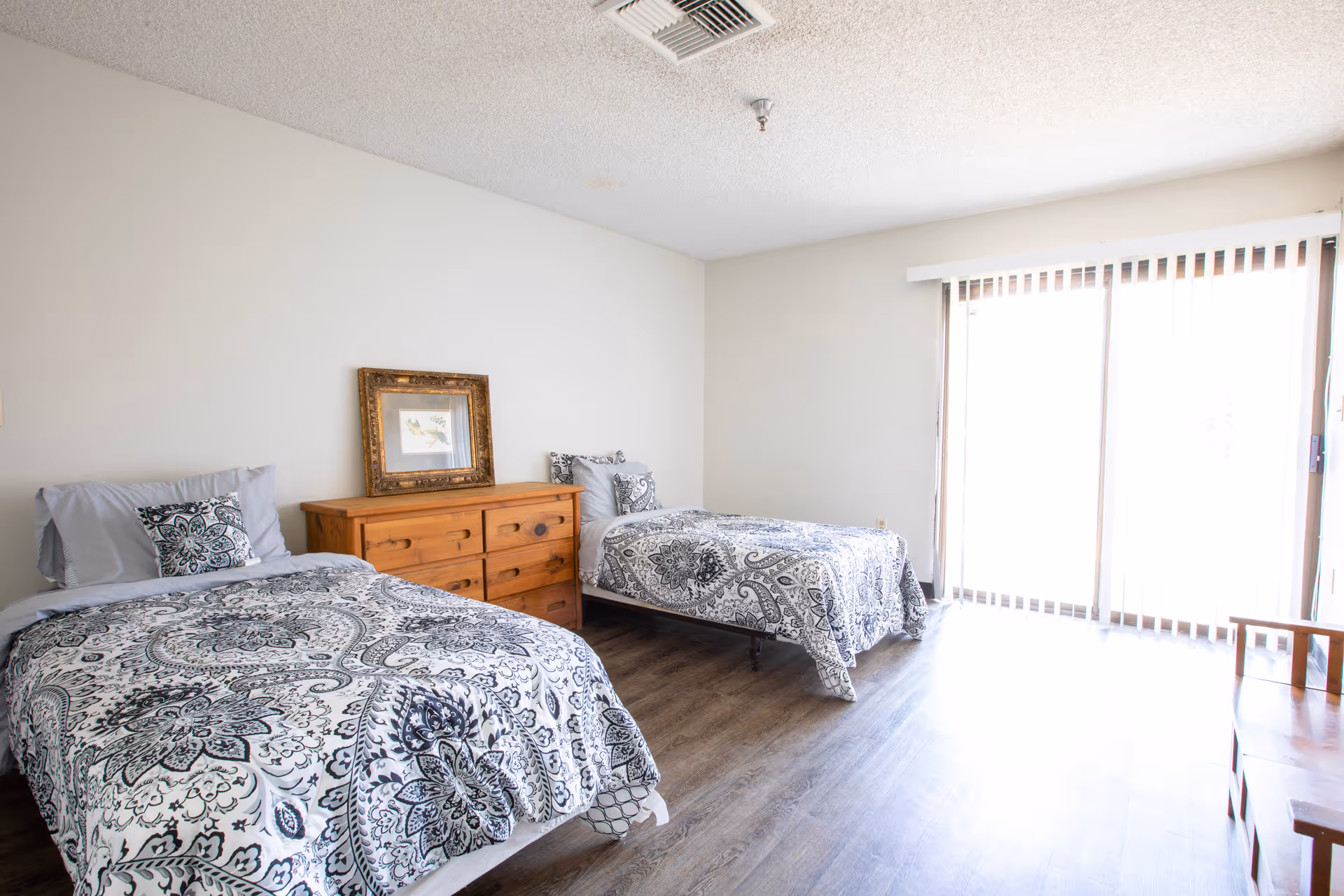 A bright bedroom with two twin beds featuring black and white patterned bedding. Between the beds is a wooden dresser with a framed picture resting on top. The room has light-colored walls, wood flooring, and a large sliding glass door with vertical blinds allowing natural light to enter.