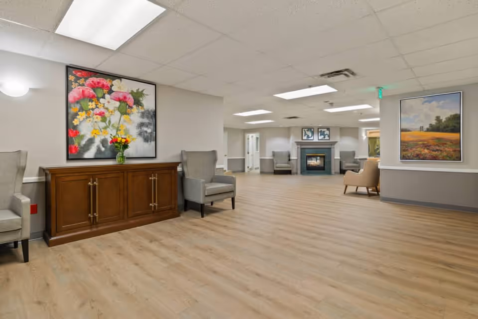 A spacious and well-lit common area in a senior living facility with light wood flooring, beige walls, and a white ceiling with recessed lighting. The room features several comfortable armchairs arranged around the space, a wooden cabinet with a vase of flowers on top, colorful floral paintings on the walls, and a fireplace with two armchairs on either side.