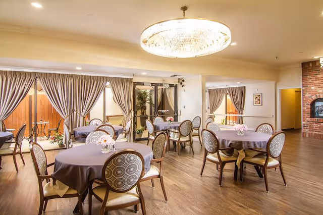 A bright dining room with round tables covered in gray tablecloths, each surrounded by patterned chairs. The room features large windows with striped curtains, a chandelier hanging from the ceiling, and a brick wall with a built-in oven or fireplace on the right side.