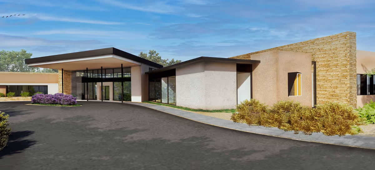 Exterior view of a modern senior living facility building with a covered entrance, large glass windows, stone and stucco walls, surrounded by landscaping including bushes and purple flowers under a blue sky.