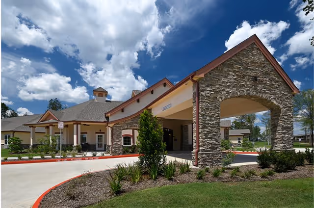 Exterior view of a senior living community building with a stone and beige facade, a covered entrance with stone pillars, landscaped greenery, and a bright blue sky with scattered clouds.