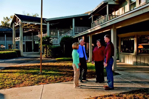 Four adults standing and talking outside a two-story building with balconies and a covered walkway, with grass and pavement around them.