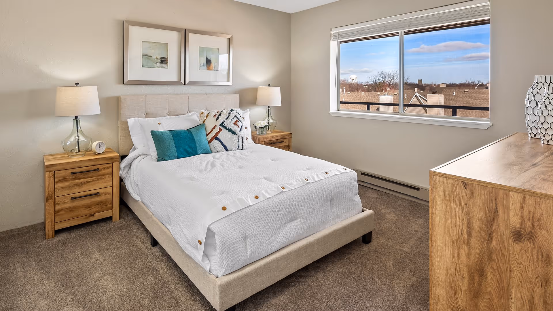 A neatly made bed with white bedding and decorative pillows in a bedroom. There are two wooden nightstands with lamps on either side of the bed. Above the bed are two framed abstract paintings. A large window shows a view of rooftops and a water tower outside. A wooden dresser with a decorative vase is on the right side of the room.