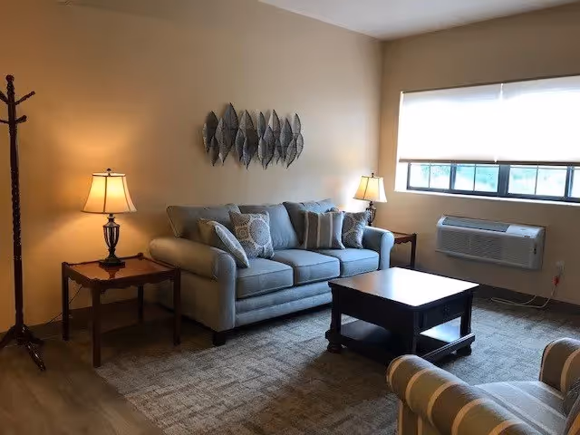 A cozy living room with a light gray sofa adorned with multiple cushions, a wooden coffee table in front, and two side tables each holding a lamp with a beige shade. A coat rack stands in the corner, and a large window with a white blind lets in natural light. The walls are painted beige, and a decorative metal wall art piece hangs above the sofa. The floor is carpeted with a patterned rug, and an air conditioning unit is installed below the window.