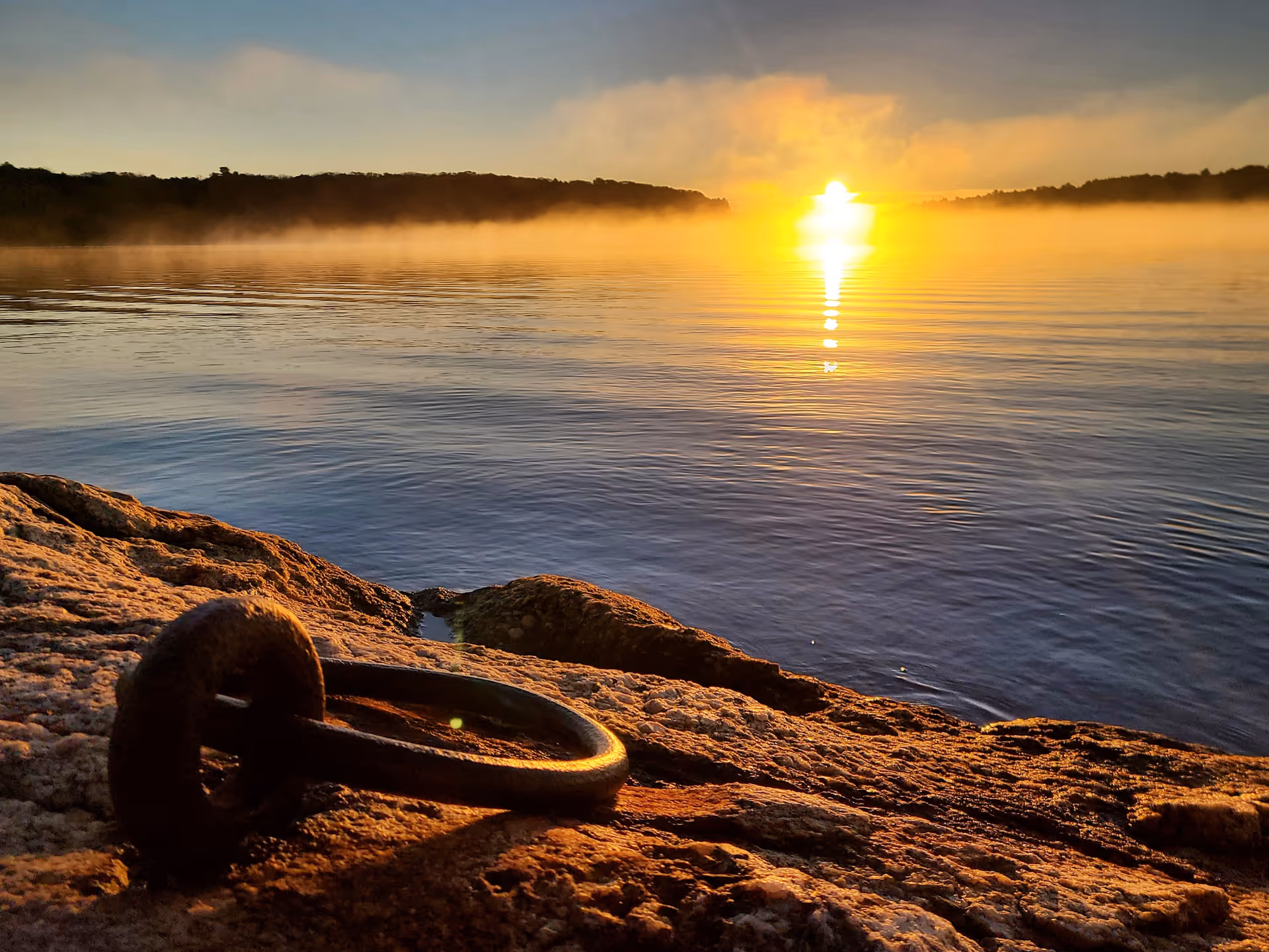 Sunrise over a calm lake with mist on the horizon and a rusted metal ring on a rocky shore in the foreground.