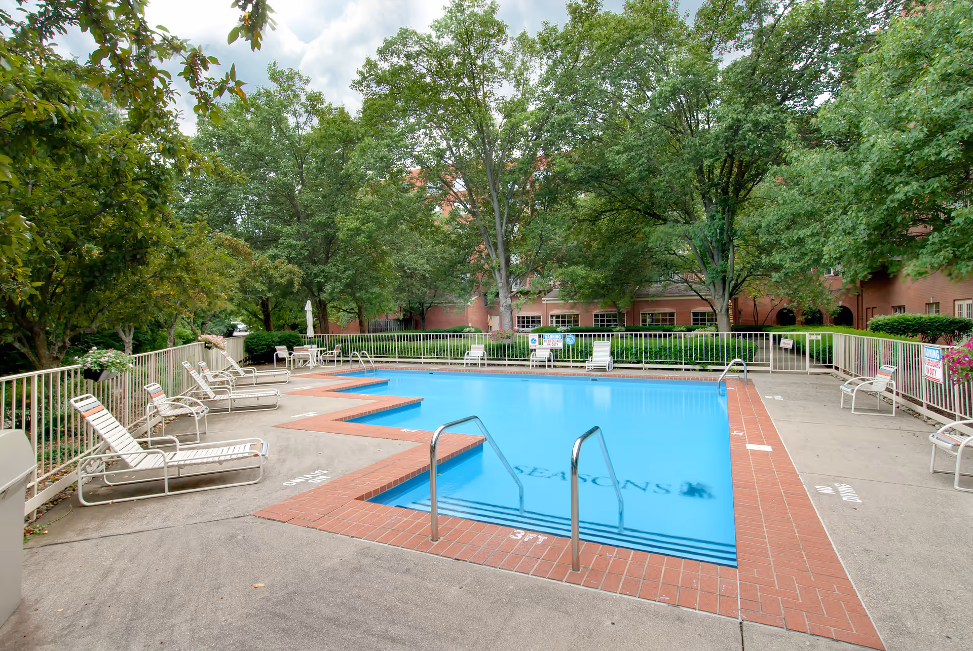 Outdoor swimming pool area with several white lounge chairs and tables with umbrellas around it, surrounded by green trees and a brick building in the background.