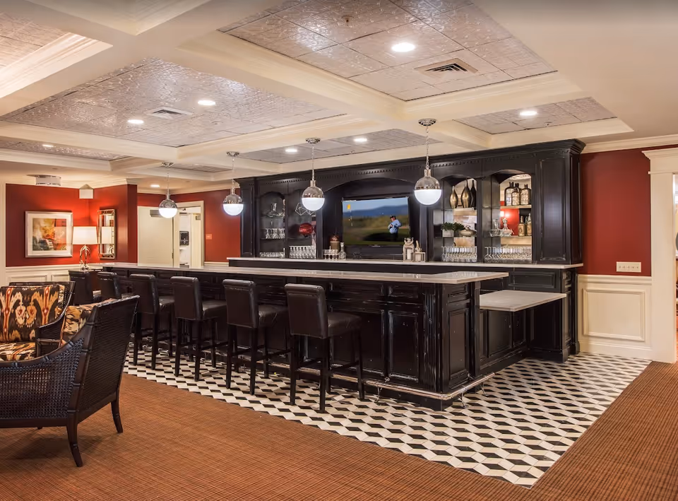A stylish senior living facility lounge area featuring a long black bar with multiple high chairs, pendant lights hanging above, and a TV mounted on the back wall. The floor has a patterned tile section near the bar and carpet elsewhere. There are decorative shelves with glassware and ornaments, and comfortable seating with patterned cushions is partially visible.