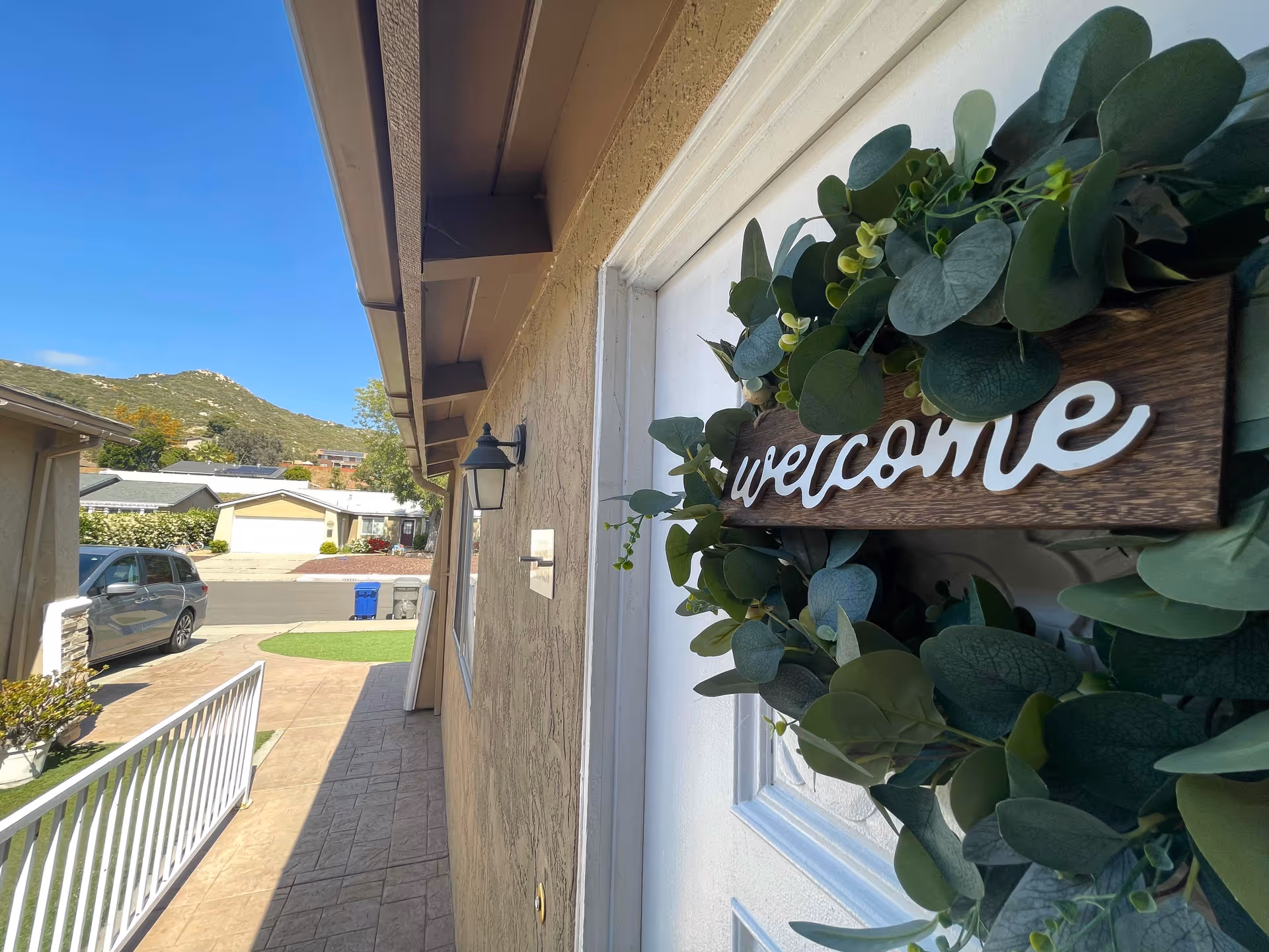 Front entry of a single-story home with a 'welcome' wreath on the door, a walkway leading to the street, a parked car, and houses with hills in the background.