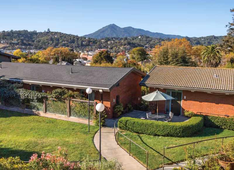Brick senior living buildings with a landscaped courtyard, patio table and umbrella, and a mountain view in the background.