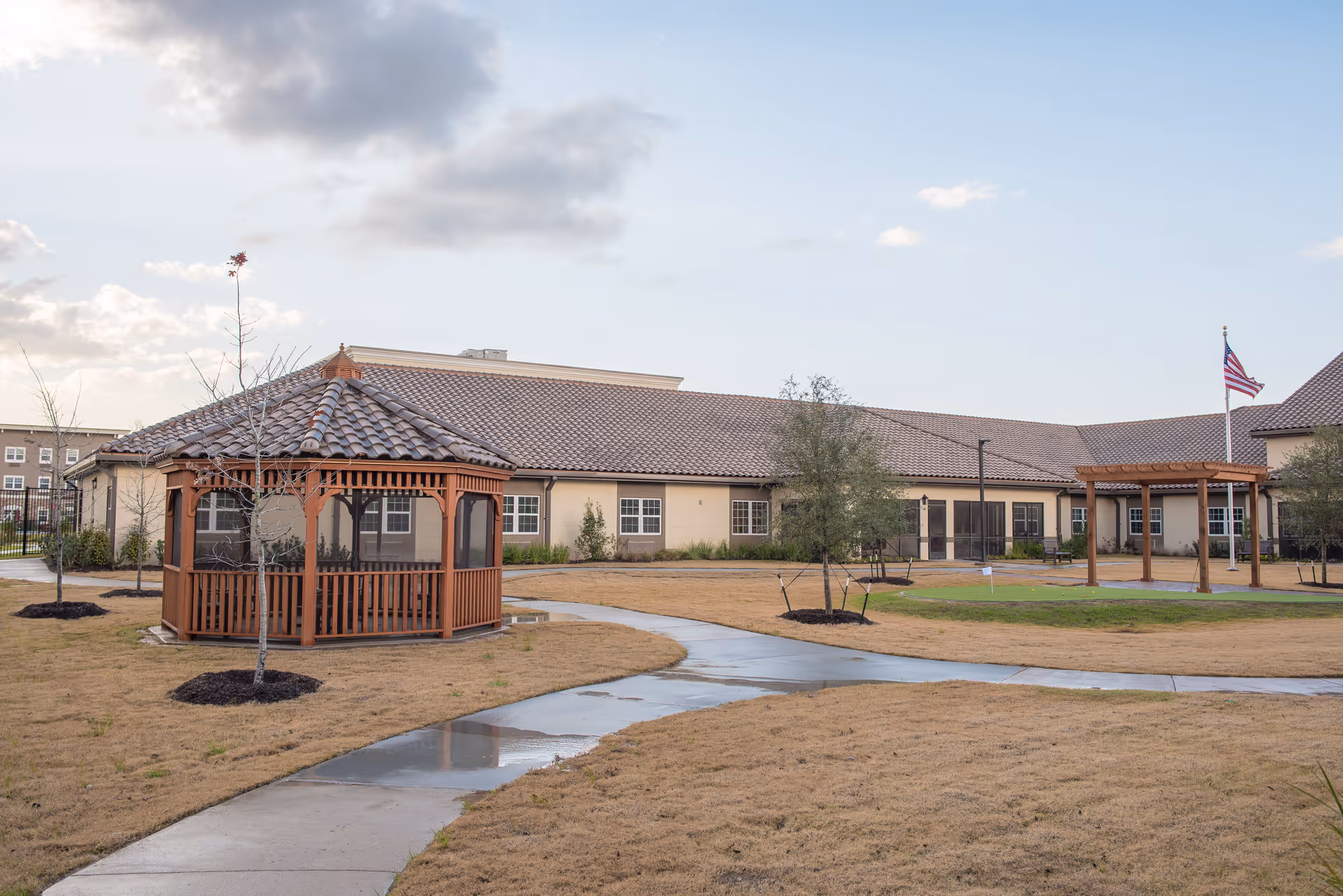 Wooden gazebo and winding concrete path in a grassy courtyard in front of a single‑story senior living building under a cloudy sky.