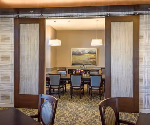 Dining room with a long table and chairs seen through large sliding wooden panels, pendant lights and a landscape painting on the far wall.