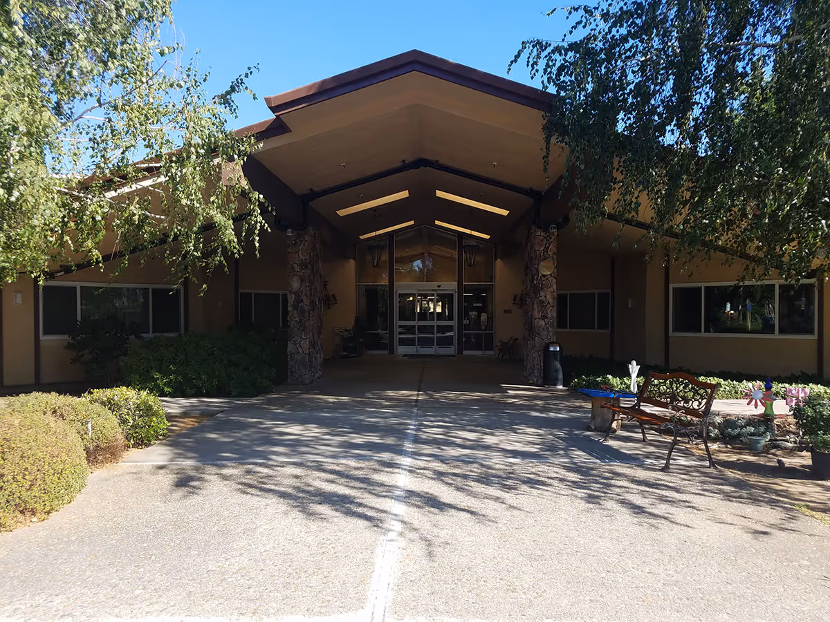 Front exterior view of a single-story building with a peaked roof entrance supported by stone pillars. There are bushes and trees on either side of the walkway leading to glass double doors. A bench and some potted plants are visible on the right side near the entrance.
