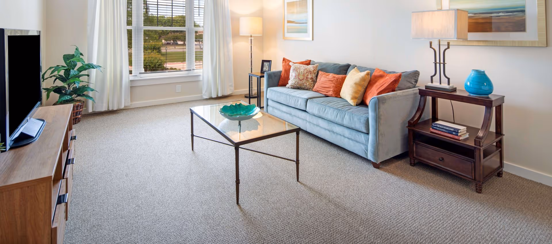 Well-lit living room with a blue sofa, colorful throw pillows, glass coffee table, TV on a wooden stand, and a large window.