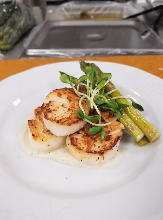 A white plate with three seared scallops garnished with green sprouts and served with three stalks of cooked asparagus on a creamy white sauce.