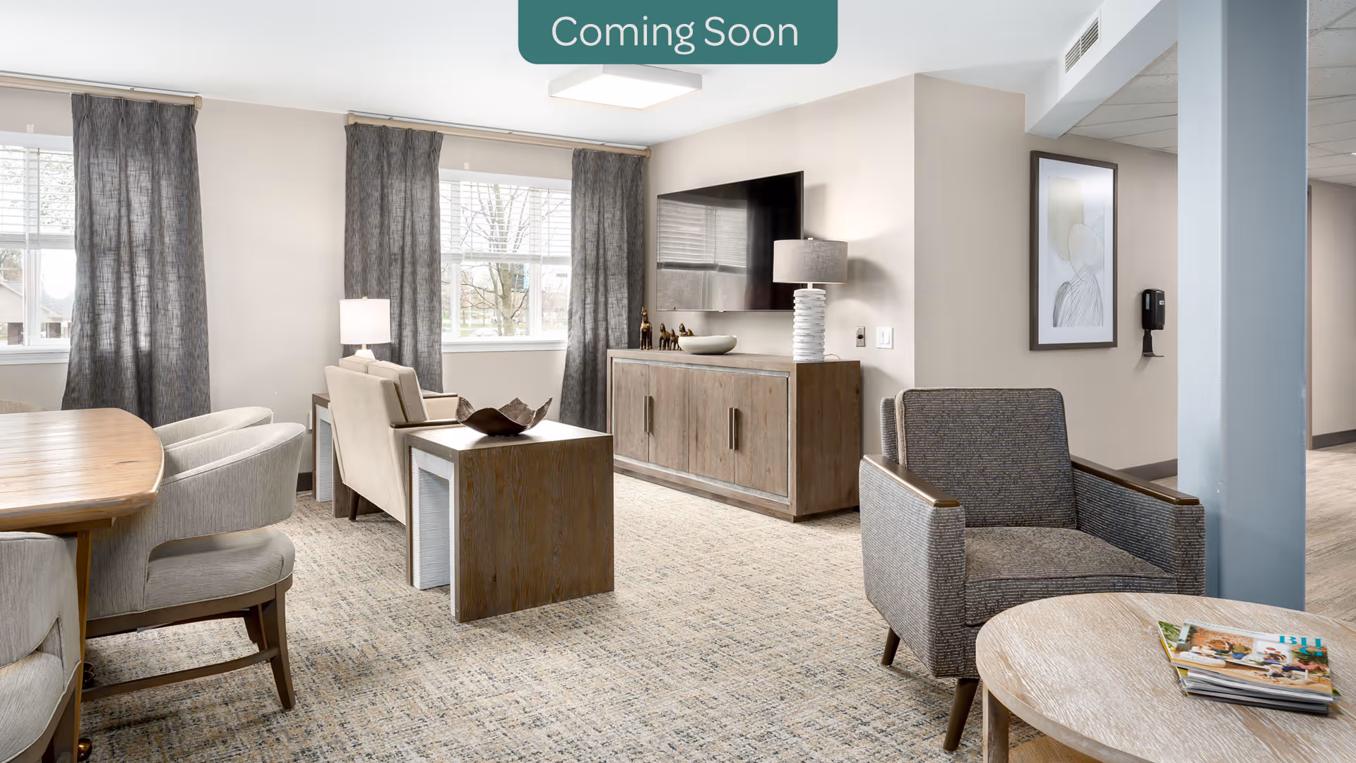 A bright and modern living room area with large windows covered by gray curtains. The room features a wooden dining table with upholstered chairs, two armchairs, a wooden side table, a wooden cabinet with a flat-screen TV mounted above it, and a round coffee table with magazines. The walls are light-colored, and there is a framed artwork and a wall-mounted hand sanitizer dispenser in the background.