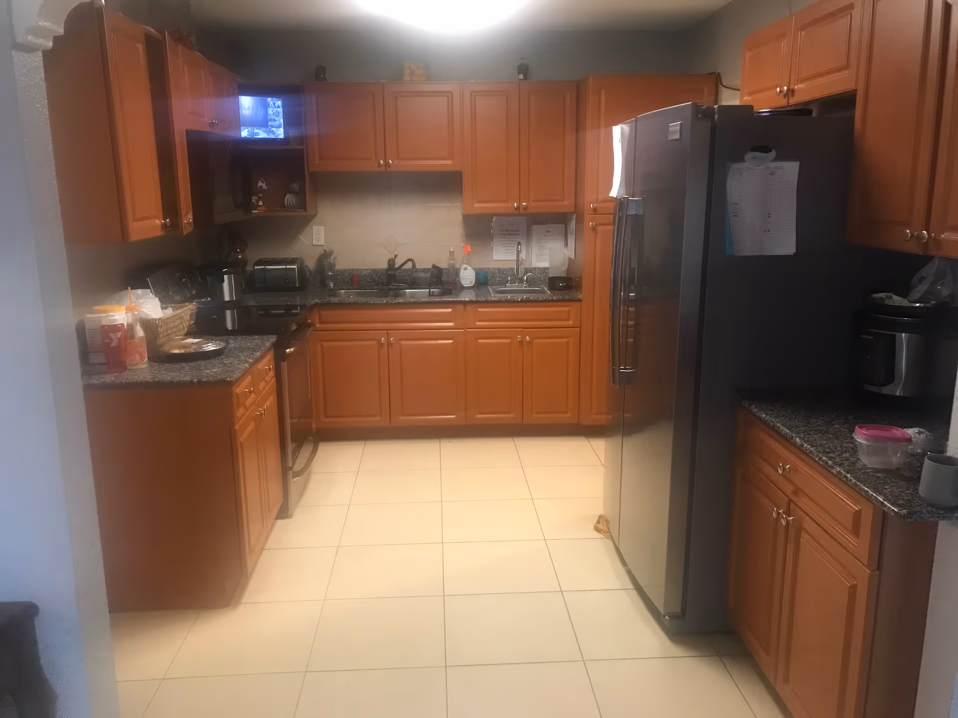 A kitchen with wooden cabinets and granite countertops. There is a stainless steel refrigerator on the right side, a stove and toaster on the left, and a double sink in the center. Various kitchen items such as a coffee maker, dish soap, and containers are placed on the countertops. The floor is tiled with light-colored tiles.