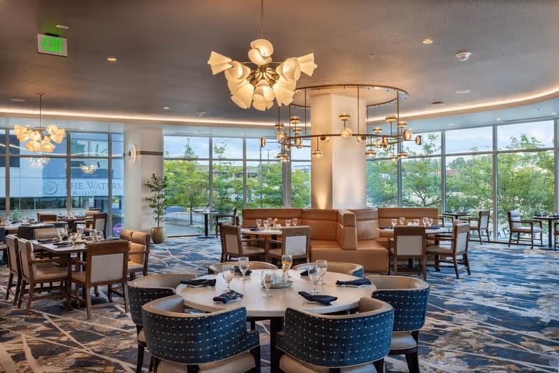 A spacious and well-lit dining room with large floor-to-ceiling windows showing greenery outside. The room features multiple dining tables set with glassware, napkins, and cutlery. There are upholstered chairs and a central seating area with a curved leather bench. Modern chandeliers hang from the ceiling, and the carpet has a blue and beige abstract pattern.
