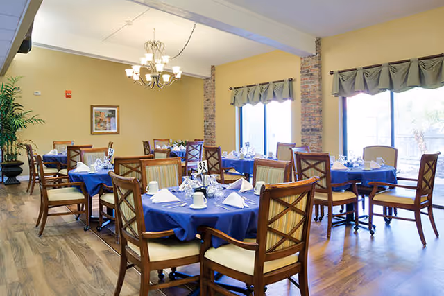 Sunlit dining room with round tables set with blue tablecloths, wooden chairs, chandeliers and large windows.