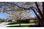 Brick senior living facility with blooming trees, green lawn, and a flag near the entrance.