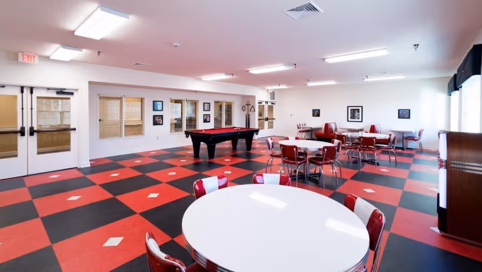 A spacious recreational room with a red and black checkered floor featuring white diamond accents. The room contains several round tables with red and white chairs, a pool table in the center, and a jukebox on the right side near large windows with blinds. The walls are decorated with framed pictures, and there are double doors on the left side.