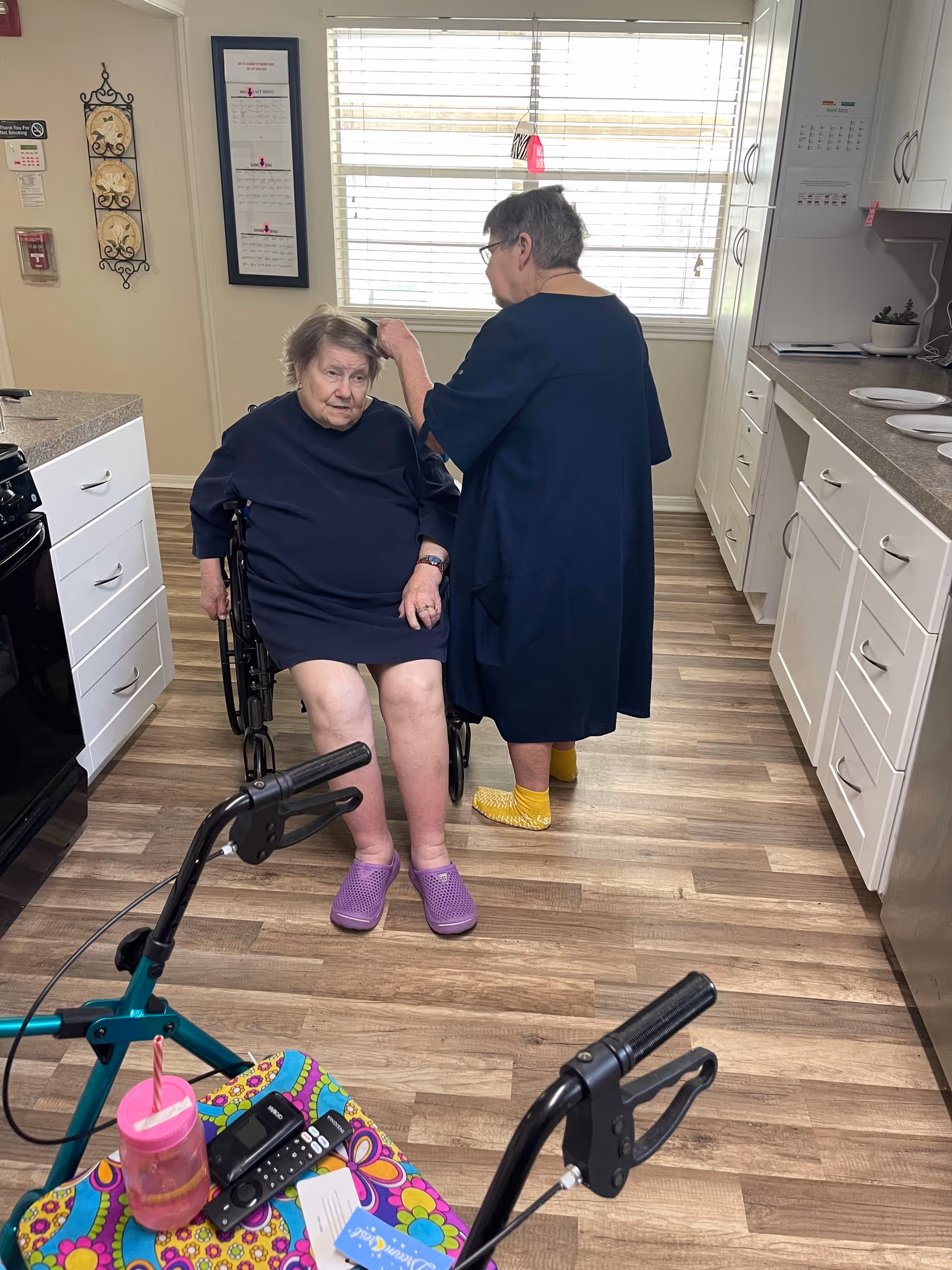 Two elderly women in a kitchen area of an assisted living facility. One woman is sitting in a wheelchair wearing purple slippers, while the other woman stands behind her, adjusting her hair and wearing yellow slippers. The kitchen has white cabinets, a window with blinds, and a colorful table with a pink cup, remote controls, and papers on it.