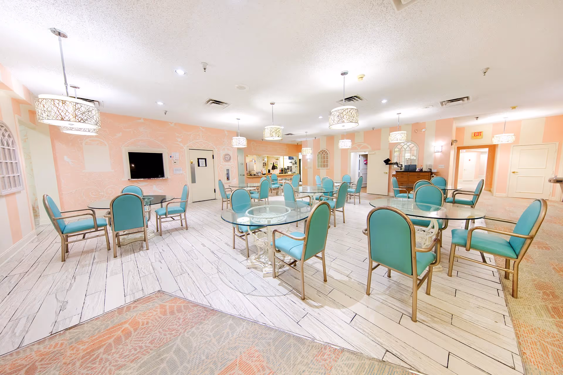 A spacious dining room with multiple round glass tables surrounded by teal cushioned chairs. The walls are painted in soft peach and pink tones with decorative patterns. Several pendant lights hang from the ceiling, illuminating the room. There is a television mounted on one wall and a serving counter in the background.