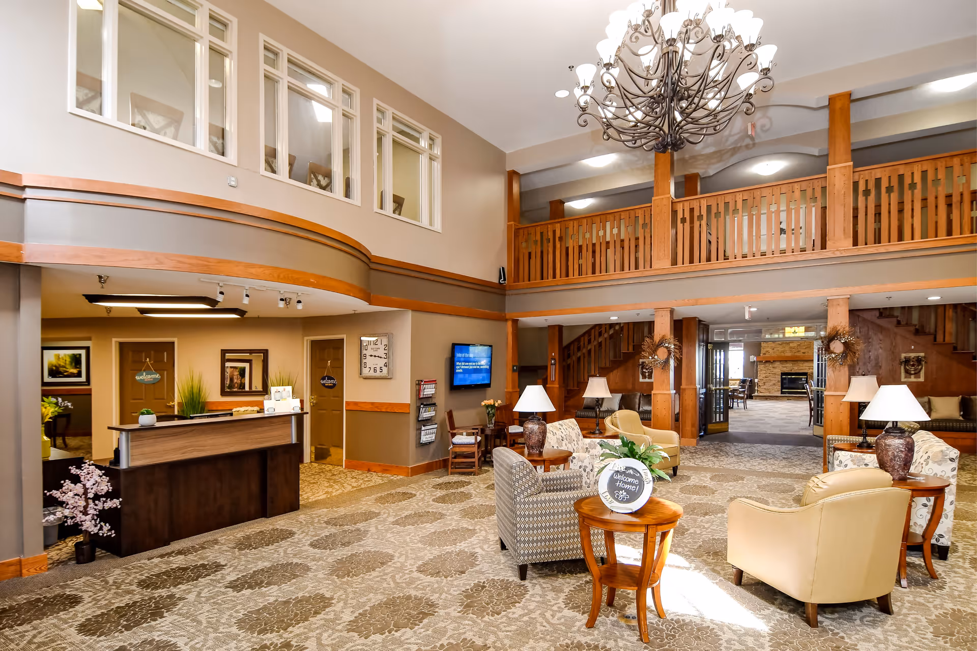 Spacious two-story senior living lobby with seating areas, a reception desk, chandelier and an upper balcony.