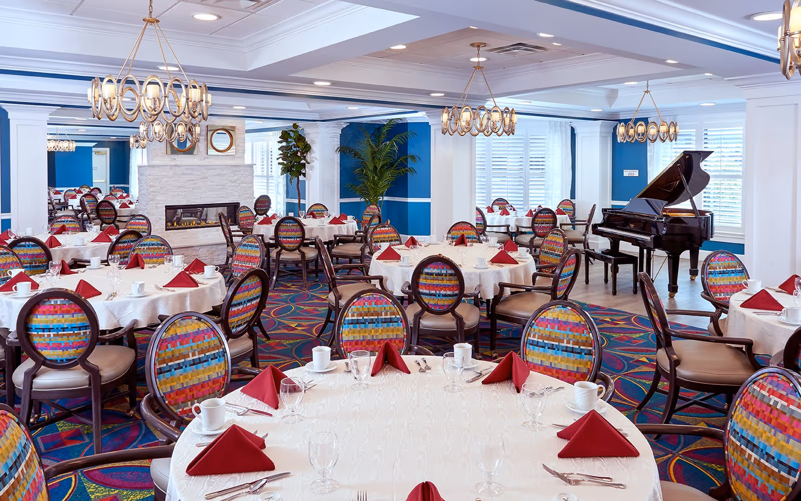 A bright and elegant dining room with multiple round tables covered in white tablecloths, each set with red folded napkins, white cups, glasses, and silverware. The chairs have colorful, patterned upholstery. The room features blue walls, white trim, a fireplace, large windows with white shutters, green plants, and a black grand piano in the corner. Modern circular chandeliers hang from the ceiling.