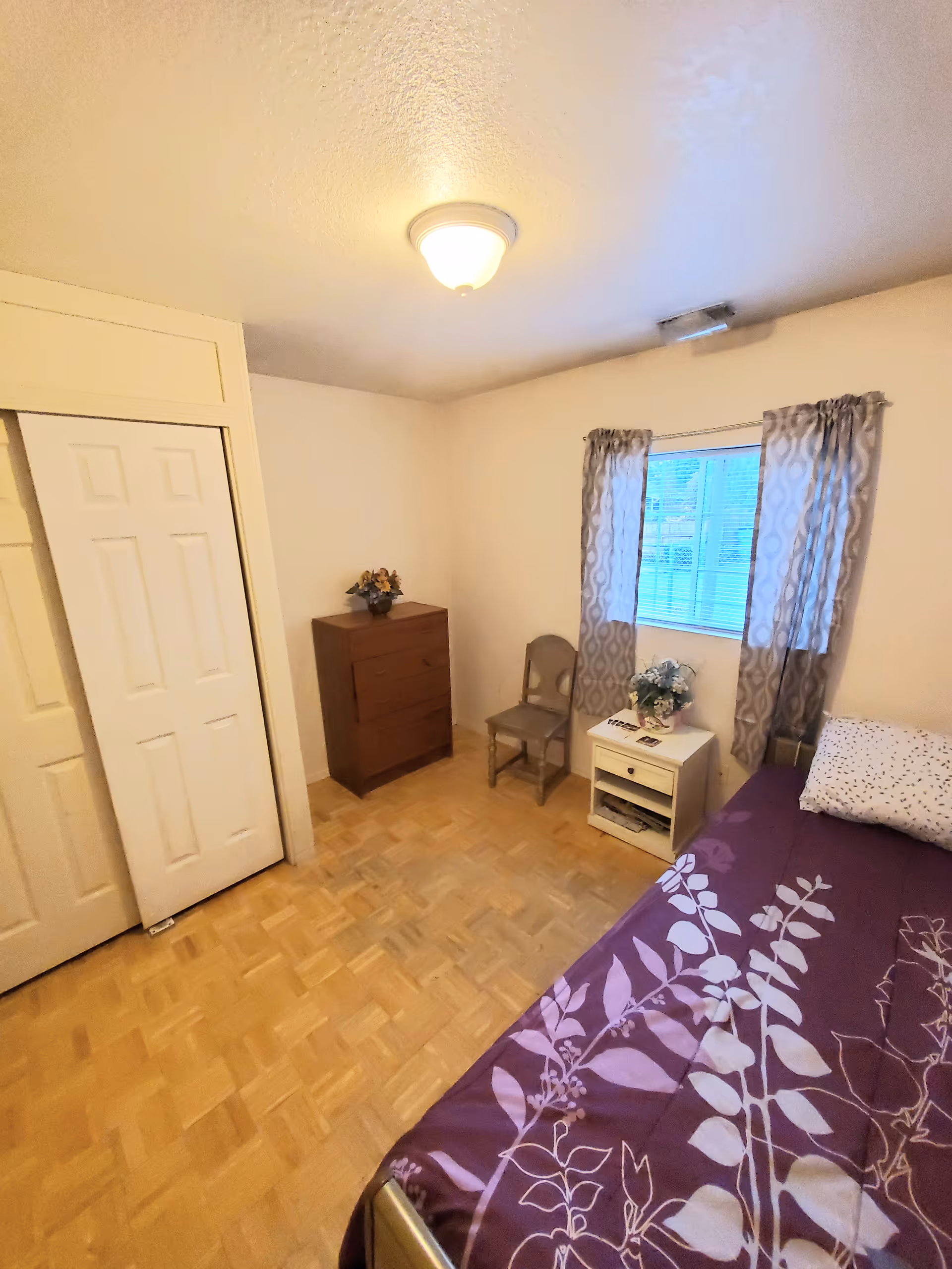 A small bedroom with a single bed covered in a purple floral bedspread, a white pillow, a wooden dresser with a flower vase on top, a wooden chair, a white nightstand with a flower arrangement, and a window with patterned curtains letting in natural light.