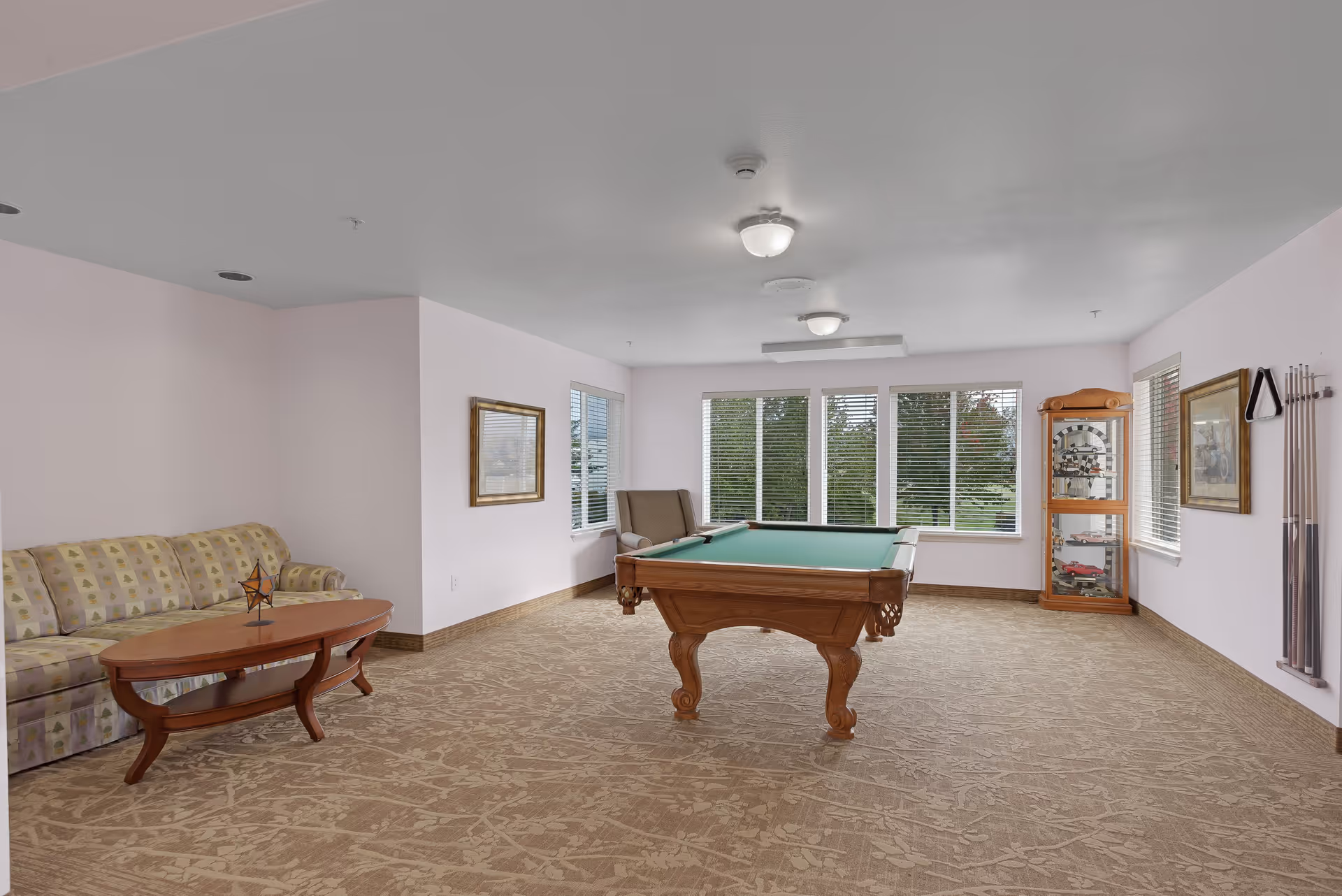 A spacious room with a pool table in the center, a patterned beige carpet, a sofa with a wooden coffee table on the left, a cushioned armchair near the windows, and a wooden display cabinet with collectibles on the right. The room has large windows with white blinds letting in natural light, light pink walls, and framed artwork on the walls.