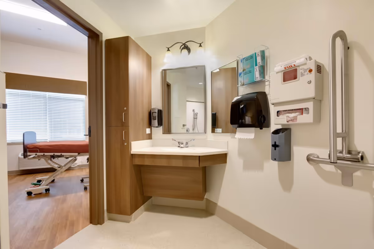 A clean and well-lit medical examination room with a wooden cabinet and a corner sink with a mirror above it. On the wall next to the sink are dispensers for paper towels, gloves, and other medical supplies. Through the open door, a medical examination table with a red cover is visible in an adjacent room with a large window covered by blinds.