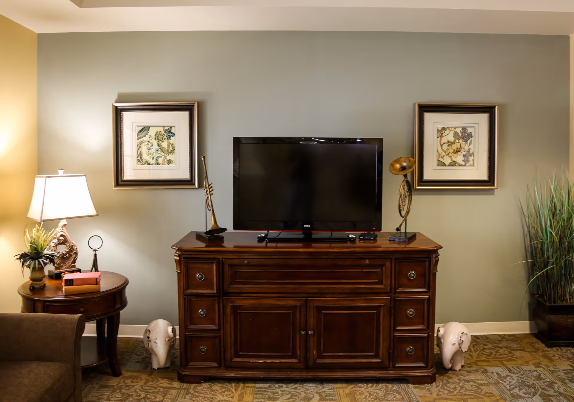 A cozy living room area featuring a wooden TV stand with a flat-screen television on top. On either side of the TV are decorative items including a brass trumpet and a vintage gramophone horn. Two framed floral artworks hang on the wall above the TV. To the left, there is a round wooden side table with a lamp, a small plant, and some books. Two white elephant figurines are placed on the floor on each side of the TV stand. A tall plant in a pot is positioned on the right side of the scene.