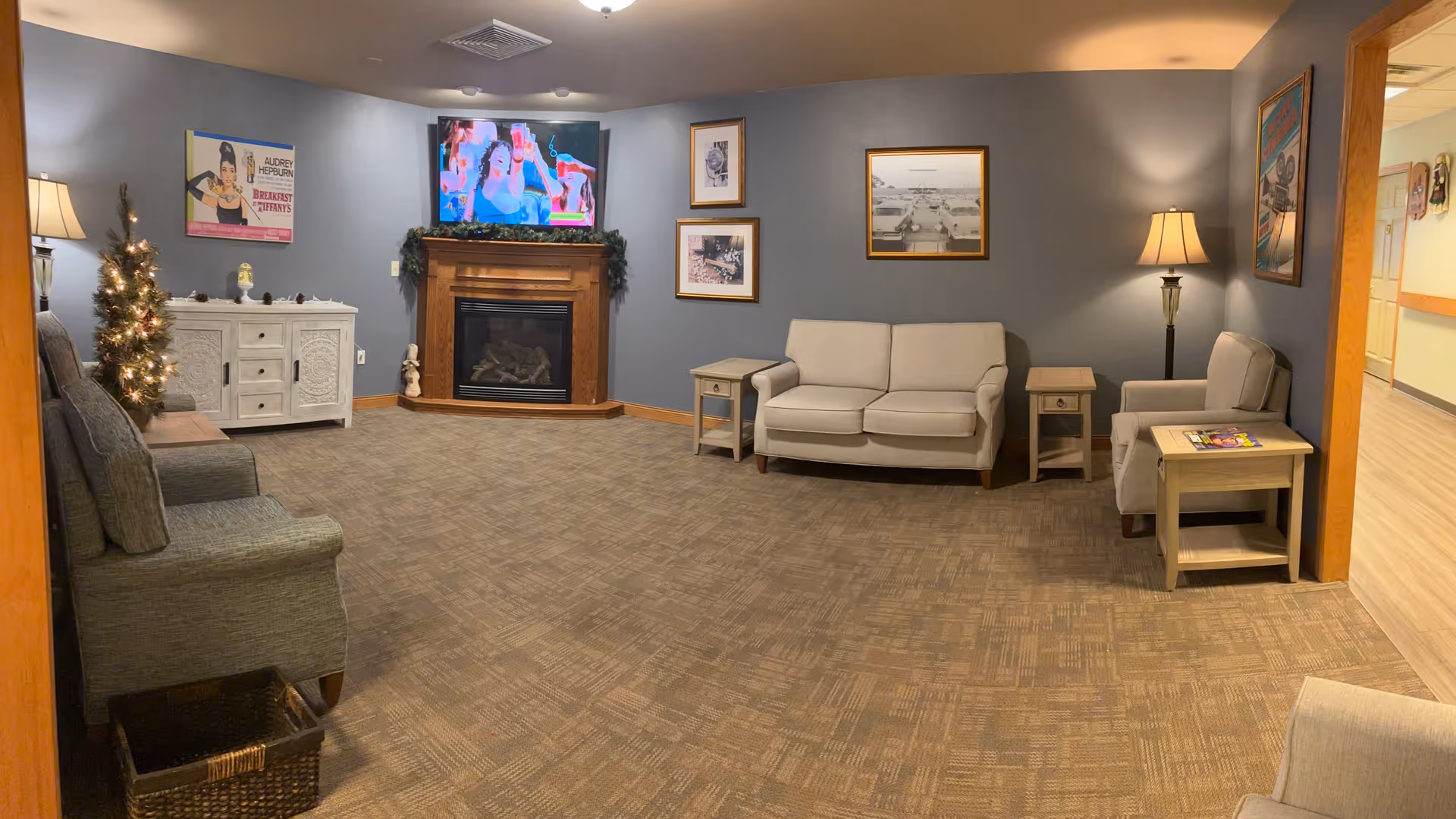 A cozy living room area with beige and gray upholstered chairs and sofas arranged around a corner fireplace with a TV mounted above it. The walls are painted gray and decorated with framed pictures and a vintage Audrey Hepburn poster. There is a small decorated Christmas tree next to a white cabinet, and two table lamps providing warm lighting. The floor is carpeted with a patterned design.