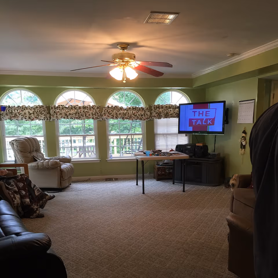 A cozy living room with green walls and beige carpet. The room features a ceiling fan with lights, three large arched windows with floral valances, a beige recliner, a black leather couch, a brown armchair, and a folding table with various items on it. A flat-screen TV mounted on the wall displays the text 'THE TALK'. There is a calendar and a small decorative hanging on the wall near the TV.